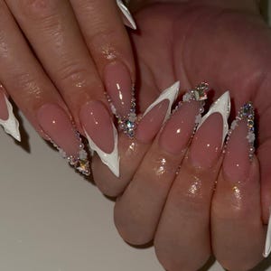 May include: Almond-shaped artificial nails with a French manicure design. The nails have a white tip and a clear, pink base. Some nails are decorated with rhinestones and small pearls for added detail.