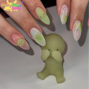 May include: A light green figurine with its hands covering its face, next to a hand with long, almond-shaped nails. The nails are painted in various designs, including a smiley face and abstract art. The logo "Berry Bop Nails" is in the upper left corner.