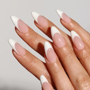 May include: Close-up of a hand with long, almond-shaped nails. Each nail features a classic French manicure design, with a white tip and a pale pink base. The nails are smooth and glossy, creating a clean and elegant look.