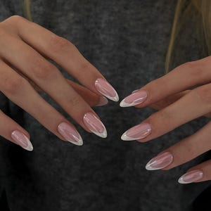 May include: Close-up of long, almond-shaped nails with a classic French manicure. The nails are painted with a sheer pink base and a crisp white tip. The hands are in front of a gray background.