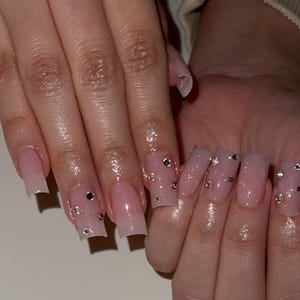 May include: Close-up of a hand with long, square-shaped acrylic nails. The nails are a light pink with glitter and small silver rhinestones. The nails have a glossy finish.