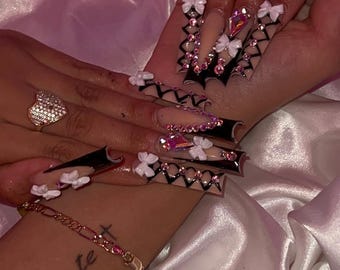 May include: Long, embellished acrylic nails with a black and nude design. The nails feature white bows, rhinestones, and a geometric pattern. A gold ring and bracelet are also visible, along with the word "delante" tattooed on the wrist.