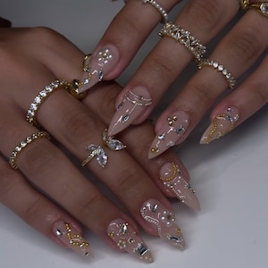 May include: Close-up of hands adorned with gold rings and long, almond-shaped nails. The nails are painted in a nude color and embellished with gold, silver, and pearl accents. The rings feature various designs, including diamonds and pearls.