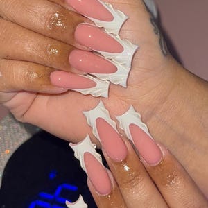 May include: Close-up of a hand with long, coffin-shaped acrylic nails. The nails feature a light pink base with a white, ruffled design around the edges. The nails are glossy and well-manicured.