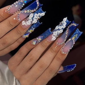 Puede incluir: Uñas artificiales largas y cuadradas con un diseño degradado, que pasa del rosa al azul. Las uñas están adornadas con pedrería, detalles de flores blancas y gemas iridiscentes, creando un aspecto glamuroso y detallado.