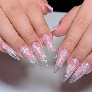 May include: Long, clear acrylic nails with a floral design. The nails feature a gradient effect, transitioning from a soft pink base to a clear tip. Delicate white floral patterns and small pearls adorn the nails, creating an elegant look.