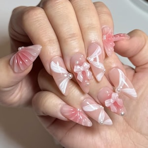 Puede incluir: Una mano que muestra uñas acrílicas largas en forma de almendra. Las uñas son de un rosa suave con diseños de lazo blanco, pequeños lazos y acentos de perlas. Algunas uñas presentan un diseño rosa degradado con una estructura en forma de abanico.