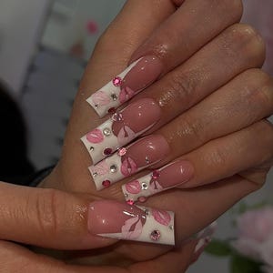 May include: Long, square-shaped artificial nails with a pink and white design. The nails feature a pink French tip, pink floral accents, and sparkling rhinestones. The design is on a white base.