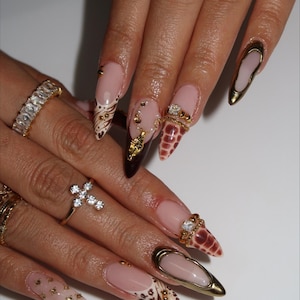 May include: A close-up of manicured nails with various designs. Some nails feature a nude base with gold accents, while others showcase animal print patterns and gold embellishments. Several gold rings with gemstones adorn the fingers.