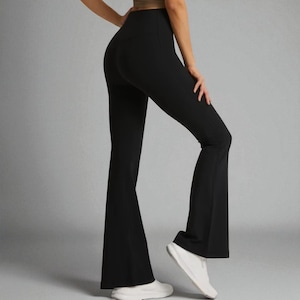 Könnte beinhalten: Schwarze Yoga-Hose mit ausgestelltem Bein. Die Hose hat eine hohe Taille und ein ausgestelltes Bein-Design. Der Stoff scheint ein glattes, dehnbares Material zu sein. Die Hose wird mit weißen Turnschuhen kombiniert. Der Hintergrund ist einfarbig grau.