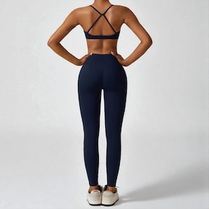May include: Navy blue athletic wear set, including a sports bra with crisscross straps and high-waisted leggings. The set is made of a smooth, stretchy material. The model is wearing white sneakers.