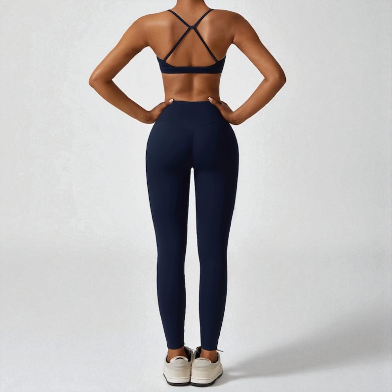 May include: Navy blue athletic wear set, including a sports bra with crisscross straps and high-waisted leggings. The set is made of a smooth, stretchy material. The model is wearing white sneakers.