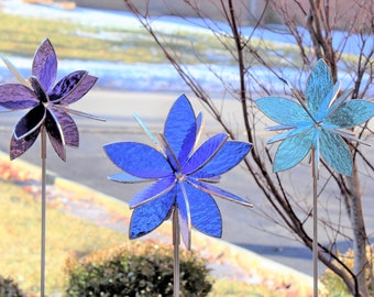 Puede incluir: Tres molinetes de viento con forma de flor de vidrio de colores con estacas de metal. Las flores son moradas, azules y verde azulado.