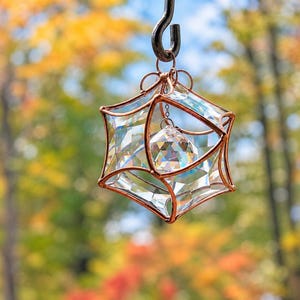 May include: A geometric suncatcher featuring a copper wire frame shaped into a star-like cage. Inside the frame, a clear, multi-faceted crystal prism hangs from a small chain, reflecting light through its angular, transparent glass surfaces.