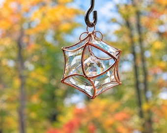 Puede incluir: Un atrapasol geométrico con un marco de alambre de cobre en forma de jaula estrellada. En el interior, un prisma de cristal transparente y multifacetado cuelga de una pequeña cadena, reflejando la luz a través de sus superficies angulares.