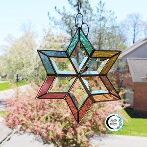 May include: A stained glass star with a geometric design in shades of blue, green, yellow, and pink. The star is hanging in a window with a blurred background of trees and a house.