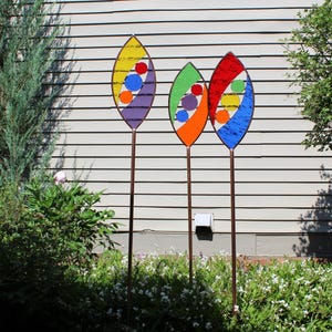 May include: Three colorful glass garden stakes with abstract designs. Each stake features a teardrop-shaped design with a mix of red, orange, yellow, blue, purple, and green glass pieces.
