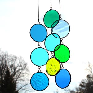 May include: A hanging stained glass mobile featuring a series of colorful, circular glass pieces in shades of blue, green, and yellow. The circles are connected by small metal chains and suspended from thin wires, creating a decorative suncatcher.