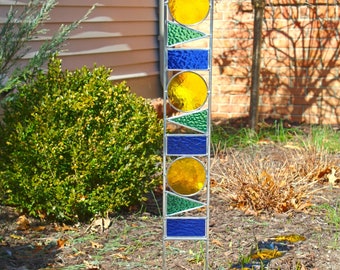 Puede incluir: Una estaca de jardín de vidrio coloreado con un diseño vertical que presenta círculos y triángulos amarillos, azules y verdes. La estaca está hecha de metal y está diseñada para colocarse en el suelo.