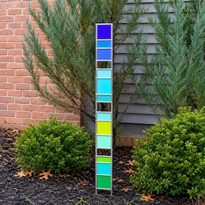 May include: A tall, vertical garden stake featuring a series of stacked, rectangular glass panels in a gradient of colors including deep blue, light blue, turquoise, lime green, and emerald green, all held within a thin, silver-toned metal frame.