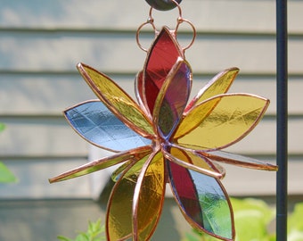 Puede incluir: Un atrapasol de vidrio coloreado en forma de flor con piezas de vidrio rojo, amarillo, azul y verde. El atrapasol está colgado de un gancho de metal negro.