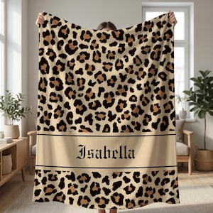 May include: A beige blanket with a leopard print pattern. The blanket has brown and black spots. A beige band with the name "Isabella" in a decorative font is centered on the blanket.