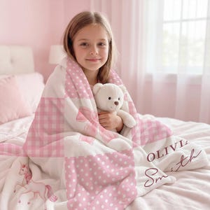 May include: A pink and white personalized blanket with a gingham and polka dot pattern. The blanket features a unicorn illustration and the name "Olivia Smith" in a cursive font. A young person is wrapped in the blanket and holding a white teddy bear.