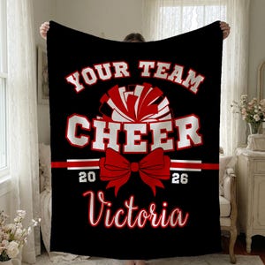 May include: Black fleece blanket with the words "YOUR TEAM CHEER" in white and red lettering. A red bow and pom-pom graphic are centered below the text. The year "2026" is printed below the bow, and the name "Victoria" is printed at the bottom.