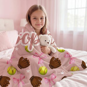 May include: A pink softball blanket featuring softballs, baseball gloves, and pink bows. The word "Softball" is written on the blanket in a script font. A young person is wrapped in the blanket, holding a white teddy bear.