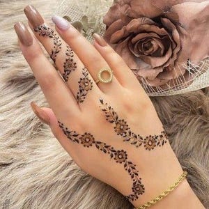 Puede incluir: Una mano adornada con intrincados diseños de henna con patrones florales y de hojas. Un anillo y una pulsera dorados añaden un toque de elegancia. Las uñas están pintadas con un color neutro.