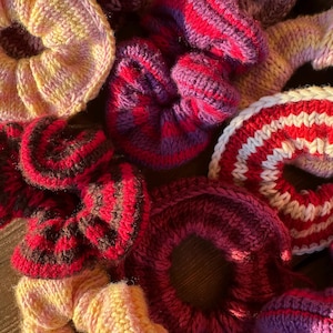 May include: A selection of handmade knitted scrunchies in various colours. The scrunchies feature shades of red, pink, purple, and yellow. Some have striped patterns, while others are solid. The scrunchies are made of soft yarn.