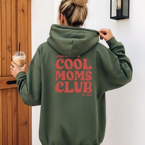 May include: Military green hoodie with the text "COOL MOMS CLUB" in red, with "EST. 2026" below. The hoodie has a drawstring hood and long sleeves. The person is holding the hood with their hand.