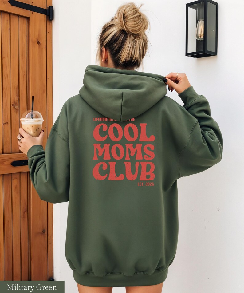 May include: Military green hoodie with the text "COOL MOMS CLUB" in red, with "EST. 2026" below. The hoodie has a drawstring hood and long sleeves. The person is holding the hood with their hand.