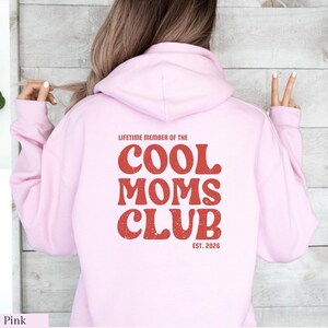 May include: A light pink hoodie with the text "LIFETIME MEMBER OF THE COOL MOMS CLUB EST. 2026" printed in red. The hoodie has a hood and long sleeves. The text is in a retro font.