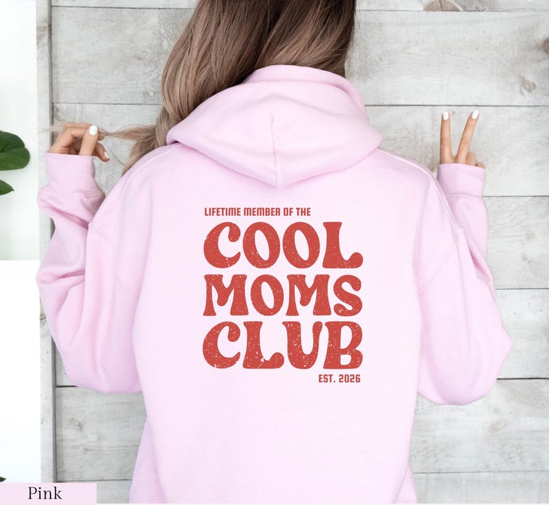 May include: A light pink hoodie with the text "LIFETIME MEMBER OF THE COOL MOMS CLUB EST. 2026" printed in red. The hoodie has a hood and long sleeves. The text is in a retro font.