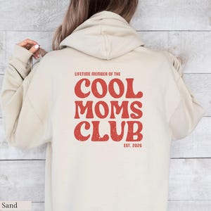 May include: Sand-colored hoodie with the text "LIFETIME MEMBER OF THE COOL MOMS CLUB EST. 2026" in red, retro-style lettering. The hoodie features a hood and long sleeves, perfect for casual wear.