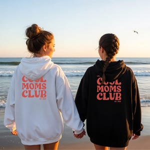 May include: Two hooded sweatshirts, one white and one black, with the text "COOL MOMS CLUB" in coral lettering on the back. The sweatshirts are worn by two people standing on a beach, holding hands, with the ocean in the background.