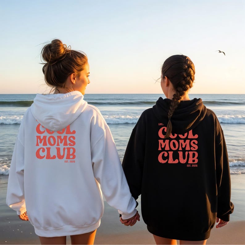 May include: Two hooded sweatshirts, one white and one black, with the text "COOL MOMS CLUB" in coral lettering on the back. The sweatshirts are worn by two people standing on a beach, holding hands, with the ocean in the background.