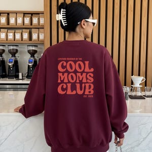 May include: Maroon sweatshirt with the text "LIFETIME MEMBER OF THE COOL MOMS CLUB EST. 2026" in a distressed orange font. The sweatshirt is a solid maroon color and has a crew neck. The image shows the back of the sweatshirt.