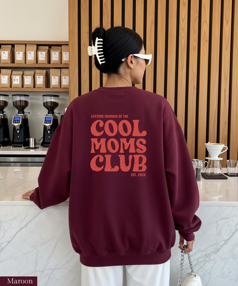 May include: Maroon sweatshirt with the text "LIFETIME MEMBER OF THE COOL MOMS CLUB EST. 2026" in a distressed orange font. The sweatshirt is a solid maroon color and has a crew neck. The image shows the back of the sweatshirt.