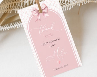 Puede incluir: Una etiqueta de regalo rosa con un lazo en la parte superior y las palabras "thank you for coming love Mila" en escritura elegante. La etiqueta tiene un borde decorativo y un pequeño corazón. La fecha "April 12, 2024" está impresa en la parte inferior.