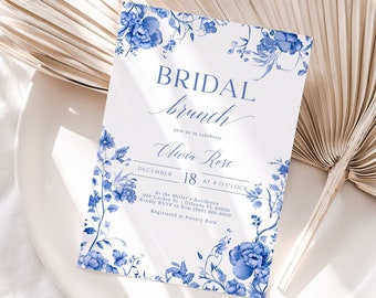May include: A white invitation card with blue floral accents and the text "BRIDAL brunch" in elegant script. The card also includes the name "Olivia Rose" and event details. The invitation is placed on a white plate with a dried palm leaf in the background.