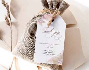 May include: A small burlap bag with a white tag attached. The tag has a floral design and the text "Thank you for showering our baby girl with love Amelia & Nick's Baby Shower November 20, 2021".