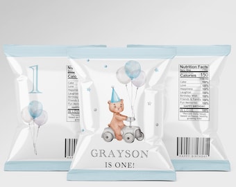 May include: A blue and white chip bag with a cartoon bear riding a toy car. The bag says "GRAYSON IS ONE!" and has a nutrition facts label.