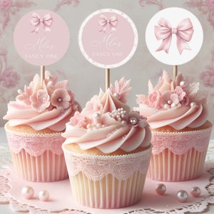 Op de afbeelding: Drie roze cupcakes versierd met roze glazuur, parelsprinkjes en roze bloemen. Elke cupcake heeft een roze en witte papieren verpakking met kant. De cupcakes zijn versierd met ronde roze en witte papieren toppers met een roze strik en de tekst "Milla's Fancy One".