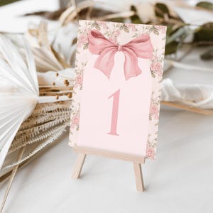 May include: A pink floral table number card with a pink bow on a wooden easel. The card has the number "1" printed on it.