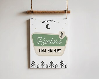 Op de afbeelding: Een rechthoekig bord met de tekst "WELCOME TO" boven een halve maan en sterren. Het bord heeft een groen en crèmekleurig gedeelte met de naam "Hunter's" en "FIRST BIRTHDAY". Onderaan een rij zwarte bomen.