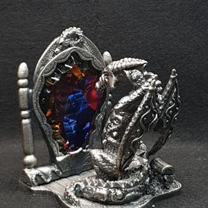 May include: A detailed silver dragon figurine with a mirror. The dragon is positioned in front of the mirror, which has a colourful, gem-like appearance. The figurine is on a small, decorative base.