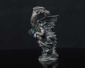Puede incluir: Una figura de dragón de color plata metálico posada sobre una base rocosa. El dragón tiene escamas detalladas, alas texturizadas y la boca abierta. Sostiene una pequeña esfera iridiscente con tonos cambiantes en azul, verde y morado.