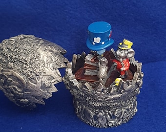 Puede incluir: Un joyero de metal plateado con un exterior texturizado de temática forestal. La tapa abierta revela una escena en miniatura con un ratón con abrigo rojo y sombrero amarillo, un sombrero de copa azul y una pila de libros.